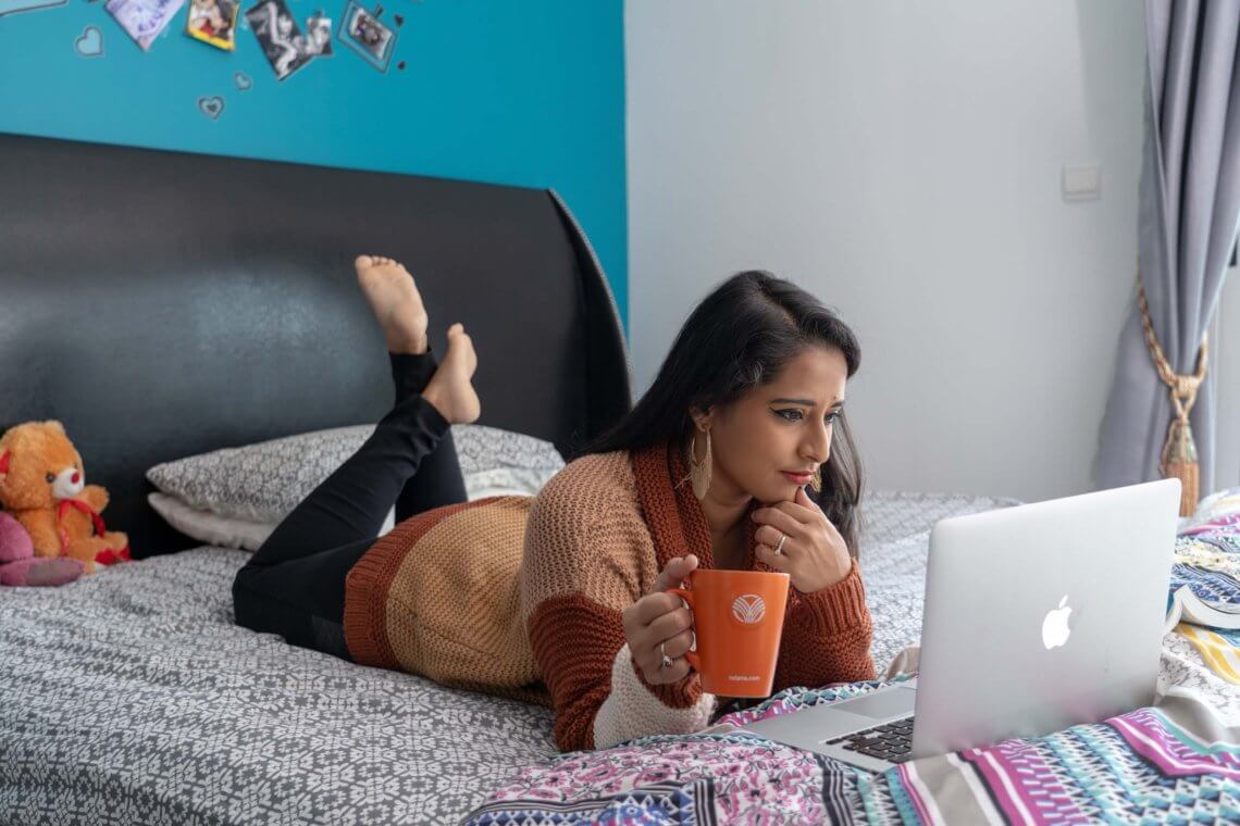WOMAN HAING COFFEE WHILE WORKING ON HER LAPTOP ON HER BED