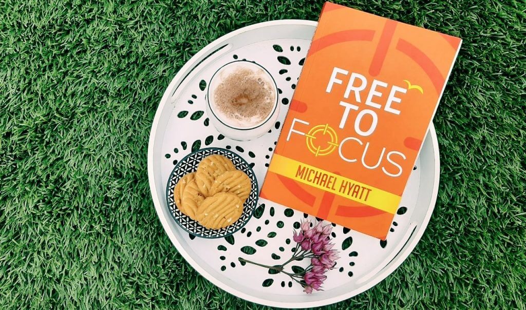 a book by michael hyatt "free to focus" is lying on a plate with some milk and cookies on grass.
